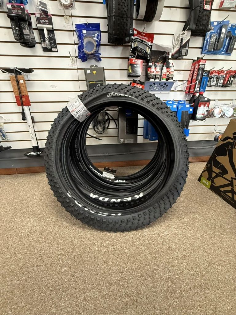 Stack of Kenda bicycle tires on shop floor in front of accessory wall display.